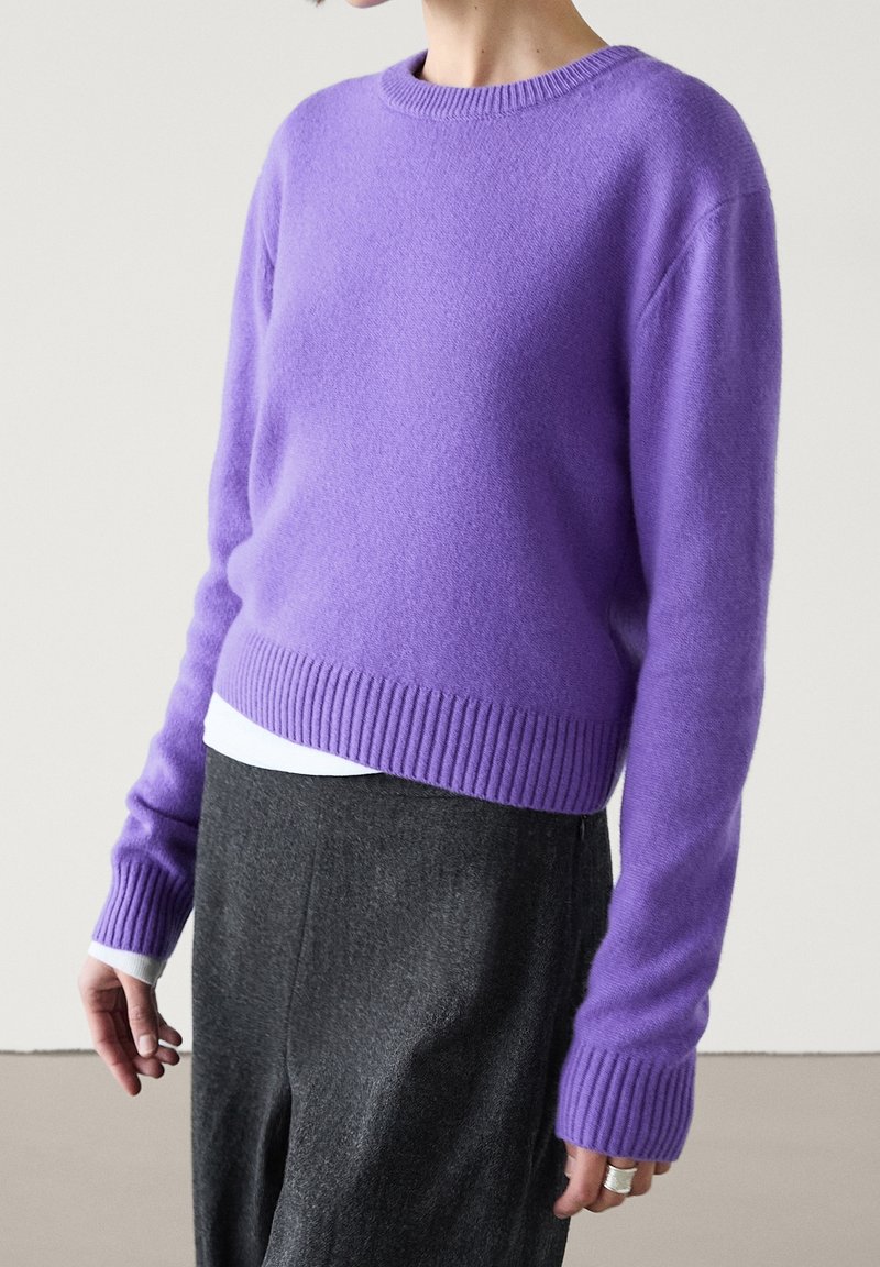 Femme portant un pull côtelé lavande à col rond manches longues sur une chemise blanche, associé à un pantalon taille haute gris foncé.