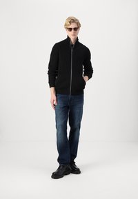 Svart ribbet strikket jakke med glidelås, sammen med blå denimjeans og svarte snørebåndsstøvler. Modellen har på seg solbriller og står med hendene i lommene.