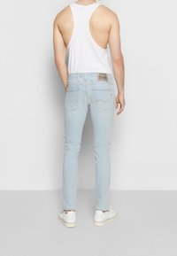 Vaqueros de mezclilla de un color azul claro con un corte ajustado, bolsillos traseros y una etiqueta tejida en la cinturilla. Combinados con una camiseta sin mangas blanca.