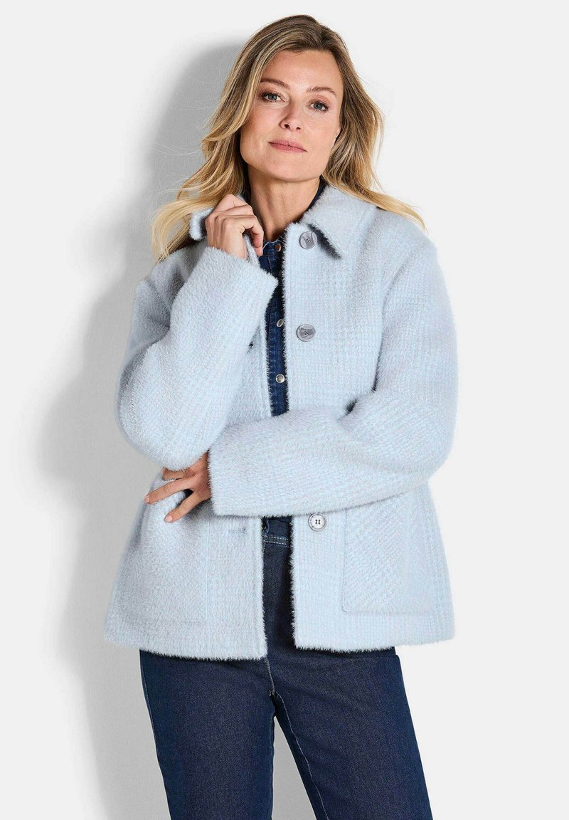 Vrouw draagt een lichtblauwe, getextureerde jas met knopen over een donkerblauw denim overhemd en donkere jeans, staande tegen een witte achtergrond.