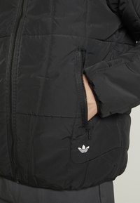 Veste matelassée noire avec fermeture éclair à l'avant, poches latérales, poignets côtelés et un petit logo blanc sur la partie gauche de la poitrine.