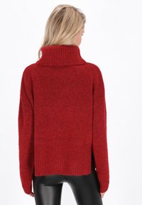 Roter, gestrickter Pullover mit hohem, geripptem Kragen, oversized Passform und strukturiertem Stoff. Verfügt über gerippte Bündchen und Saum sowie seitliche Schlitze.