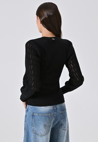 Schwarzer Strickpullover mit strukturierten, gitterförmigen langen Ärmeln. Einfarbiger Rücken und gerippte Bündchen, kombiniert mit hellen Jeans.