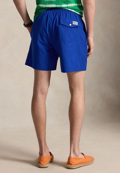 Άνδρας με μπλε casual σορτς με πίσω τσέπη και πορτοκαλί παπούτσια slip-on, στέκεται μπροστά σε ουδέτερο φόντο.