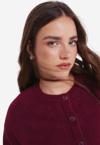 Burgundská pletená vesta s kulatým výstřihem, se dvěma tmavými knoflíky vpředu a měkkou texturou. Modelka má dlouhé hnědé vlasy.