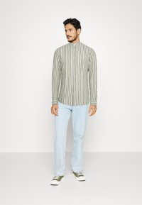 Camicia button-up in verde oliva chiaro con righe bianche verticali, maniche lunghe, colletto alla mandarino e taschino sul petto. Abbinare a jeans azzurri.