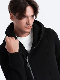 Zwarte zip-up hoodie met een grote capuchon, gemaakt van een gladde stof en voorzien van een zilveren rits, gedragen over een wit shirt. Arm is omhoog geheven.