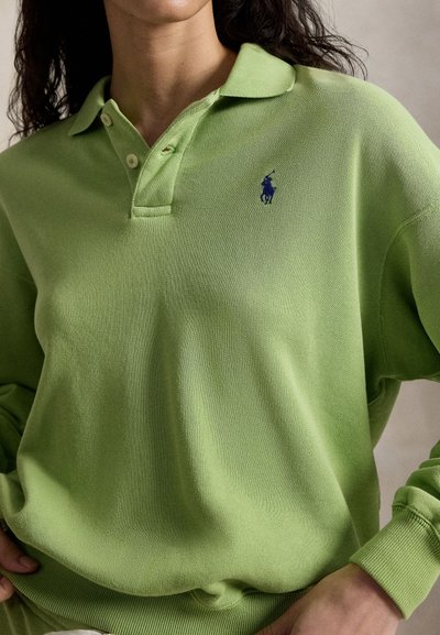 Kvinde iført en lysegrøn langærmet poloshirt med en mørkeblå broderet hest og rytter-logo på brystet.