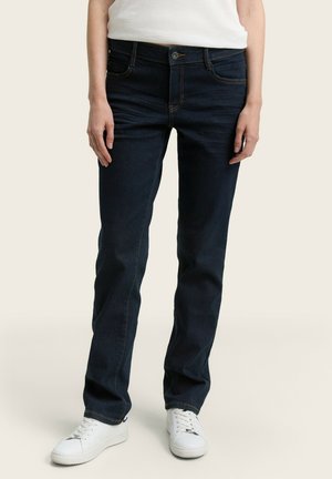 Personne portant un jean droit bleu foncé et des baskets blanches, debout devant un fond clair uni.