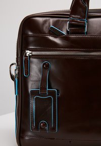 Borsa in pelle marrone con accenti blu, una tasca con zip frontale e un portatag personalizzabile. Texture liscia con dettagli di cucitura in evidenza.