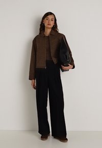 Casaco curto castanho texturizado sobre uma blusa ajustada escura. Calças pretas largas de cintura alta. Saco preto na mão, sapatos castanhos.