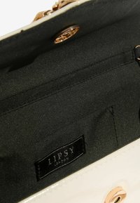 Interiorul unei genți de mână din piele lacuită crem, căptușit cu negru. Dispune de un buzunar cu fermoar și o etichetă Lipsy London. Accesorii din metal auriu.