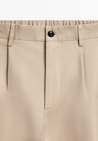 Pantaloni beige chiaro su misura con fronte piatto, cintura elastica, un bottone nero e tessuto leggermente strutturato.