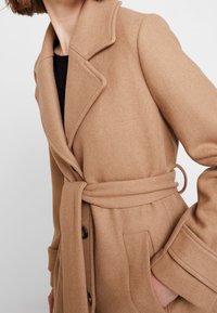 Manteau en mélange de laine camel avec taille ceinturée, col à revers et poches plaquées. Comprend des accents de boutons et une finition douce et texturée.