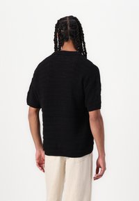 Cămașă scurtă din tricot negru cu un model texturat, guler rotund și croială relaxată, asortată cu pantaloni lăsați de culoare deschisă.