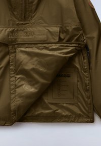 Φούτερ σε ελιάς πράσινο με μερικώς ξεκούμπωτο μπροστινό μέρος, που δείχνει την εσωτερική επένδυση με κυκλικά λογότυπα και μια ραμμένη ετικέτα με λεπτομέρειες για το ύφασμα και τη φροντίδα.