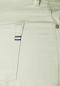 Pantalons vert clair avec une texture lisse et une petite poche avant présentant une accents rayés en noir et blanc. Détail de rivet en métal visible.