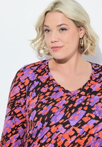 V-kaulus paita, jossa on musta kangas, jossa on eläväinen punaisen ja violetin epäsäännöllisten muotojen kuvio. Sileä pinta lyhyillä hihoilla.