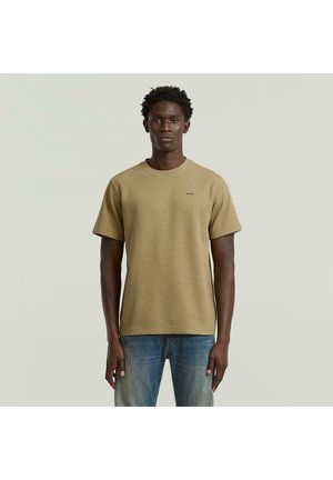 Ung mand stående iført en beige tekstureret kortærmet skjorte og falmede blå jeans, foran en ensartet lys baggrund.