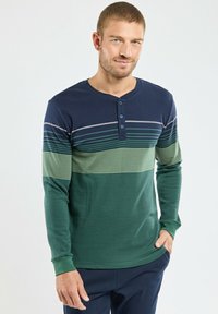 Homme portant une chemise henley à manches longues rayée en marine, vert et jaune clair, se tenant avec une main dans sa poche devant un fond uni.