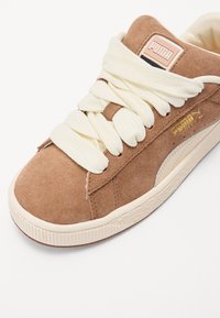Brown suede sneaker with cream laces, white midsole, and subtle gold logo. Rounded toe and textured outsole for grip. Classic design.