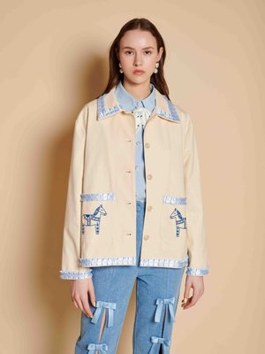 Jonge vrouw draagt een beige jas met blauwe paardenprints en ruches, lichtblauw overhemd en spijkerbroek met blauwe strikken.