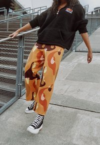 Sudadera de forro polar negra con media cremallera y bolsillo frontal, combinada con pantalones anchos de patrón naranja y marrón, y zapatillas de plataforma negras.