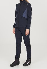 Giacca sportiva blu navy con colletto alto, tasca laterale con zip e pannello azzurro chiaro; abbinata a pantaloni blu navy aderenti e stivali neri.
