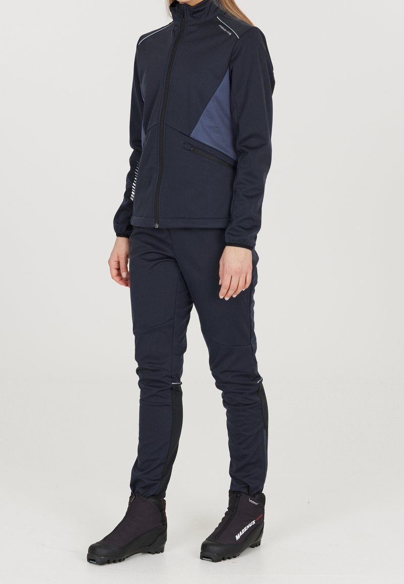 Giacca sportiva blu navy con colletto alto, tasca laterale con zip e pannello azzurro chiaro; abbinata a pantaloni blu navy aderenti e stivali neri.