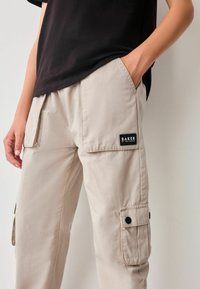 Beige Cargo-Hosen aus leichtem Stoff, mit Seitentaschen, einem Patch mit dem Label "BAKER" und einer lockeren Passform.