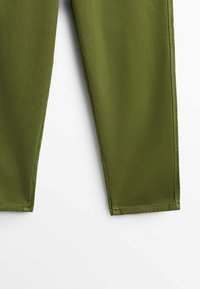 Partie inférieure d’un pantalon vert olive droit sur fond blanc, montrant les coutures de l’ourlet et la texture du tissu.
