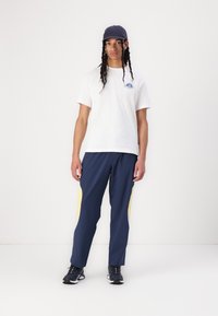 Hvit bomulls t-skjorte med blå grafisk design, kombinert med marineblå treningsbukser med gule sideakenter og svarte joggesko.