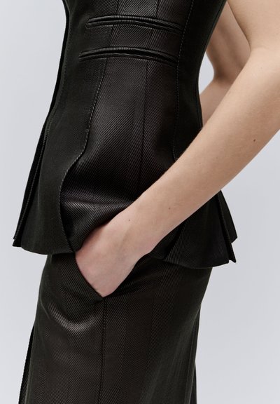 Zwart slim-fit blazer met visgraatstructuur en bijpassende rok, met hand nonchalant in de zak van de rok tegen een lichte achtergrond.