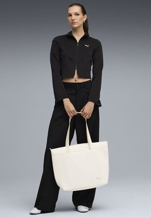 Bolsa de tela blanca con dos asas y un logo cosido, sostenida por una persona que lleva una chaqueta negra corta y pantalones negros.
