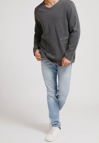 Grau langärmliges V-Ausschnitt-Shirt mit einem subtilen grafischen Detail an der Seite, kombiniert mit hellblauen Jeans und weißen Sneakers.
