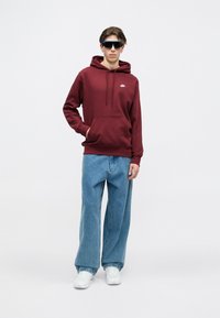 Bordová mikina s predným vreckom a šnúrkou, skombinovaná s voľnými modrými džínsami. Biele tenisky a slnečné okuliare dopĺňajú outfit.
