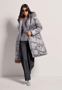 Vrouw die loopt in een grijze pufferjas met bontkraag, grijze trui, zwarte broek, grijze enkellaarsjes, zonnebril en een bruine tas.