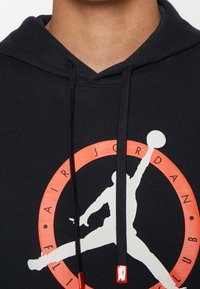 Sweat à capuche noir avec le logo Air Jordan montrant une silhouette blanche d'un joueur de basketball sautant, entourée d'une bordure circulaire rouge avec du texte.