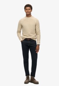 Suéter de punto beige con cuello redondo, jeans azul marino y botas de cordones marrones. El suéter tiene un patrón texturizado y puños acanalados.