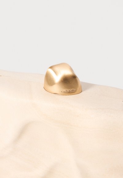 Objet en laiton en forme de roche avec une surface lisse et réfléchissante ; positionné sur une base texturée beige clair. Gravé de "cult gaia."