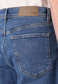 Os jeans de ganga azul apresentam um patch de couro na cintura com a inscrição "SELECTED HOMME" em relevo. Os bolsos traseiros são cosidos com linha em contraste.