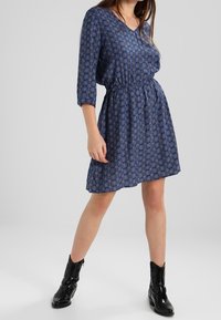 Robe bleue à motifs avec des manches trois-quarts, taille cintrée et décolleté en V, portée avec des bottines noires. Texture de tissu lisse.