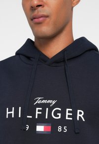 Σκούφος φούτερ ναυτικού χρώματος με κορδόνι, μπροστινό λογότυπο "Tommy HILFIGER," με έγχρωμο σχέδιο σημαίας από κάτω. Λείος υφασμάτινος ιστός.