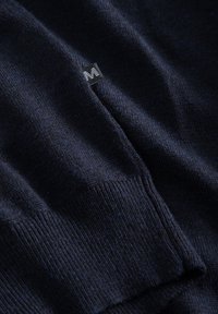 Ciemnogranatowy sweter wykonany z dzianiny o gładkiej fakturze, z małą czarną metką z białym logo po boku.