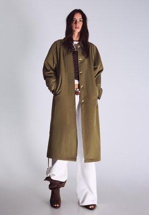 Hosszú sötét hajú nő olívazöld, gombos hosszú kabátot, fehér bő szárú nadrágot, barna nyitottorrú csizmát visel, egyszerű háttér előtt áll.