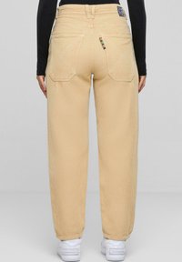 Pantaloni in corduroy beige con una vestibilità relaxed, caratterizzati da due tasche posteriori e un accento colorato sulla tasca destra. Sneakers bianche completano il look.