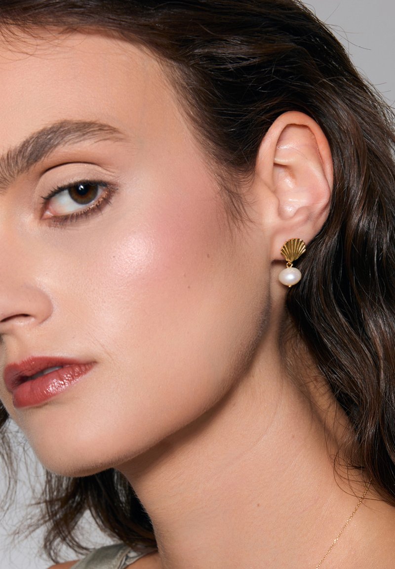 Złote kolczyki w kształcie muszli z białym akcentem w postaci perły. Kolczyki nosi modelka z włosami średniej długości.