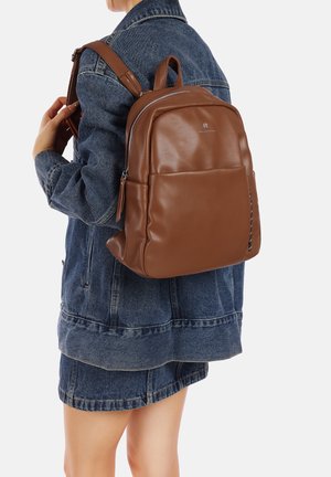 Zaino in pelle marrone con texture liscia e forma arrotondata, dotato di una tasca frontale e spallacci regolabili, indossato sopra una giacca di jeans.