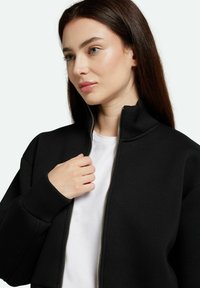 Černá zipová mikina s vysokým límcem, vyrobená z měkké tkaniny. Obsahuje stříbrný zip a bílý tričko pod ní. Hladká textura.