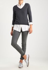 Maglione a V blu navy indossato sopra una camicia bianca con bottoni, abbinato a pantaloni grigi aderenti e scarpe stringate argento.